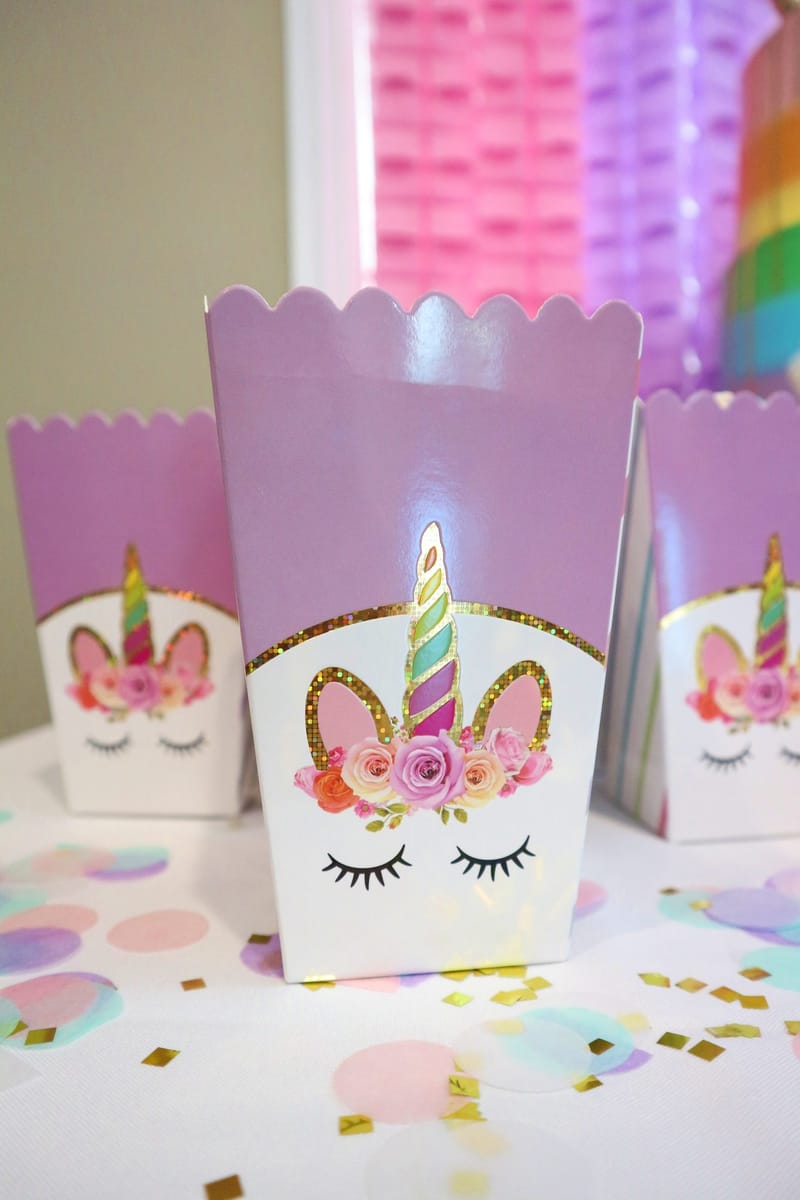 Purple and white popcorn boxes printed with a unicorn face and a gold horn, on scattered confetti