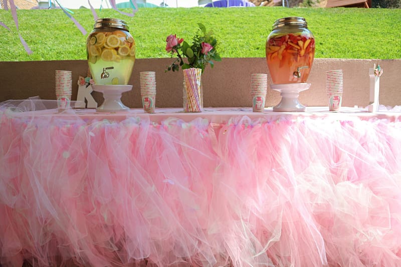 Table skirted in pink tulle with lemon and orange drink dispensers, paper cups and roses, a grassy slope behind