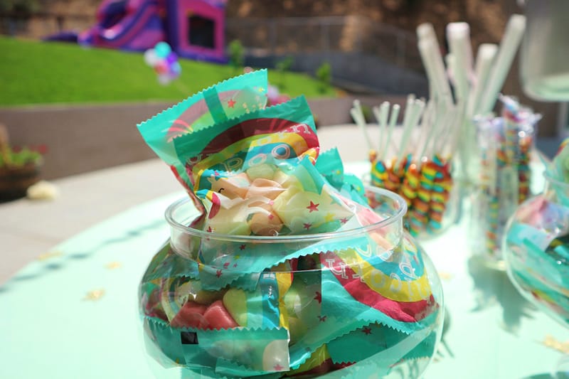 Glass bowl of wrapped rainbow gummy candy on a mint table, a bounce house in the distance