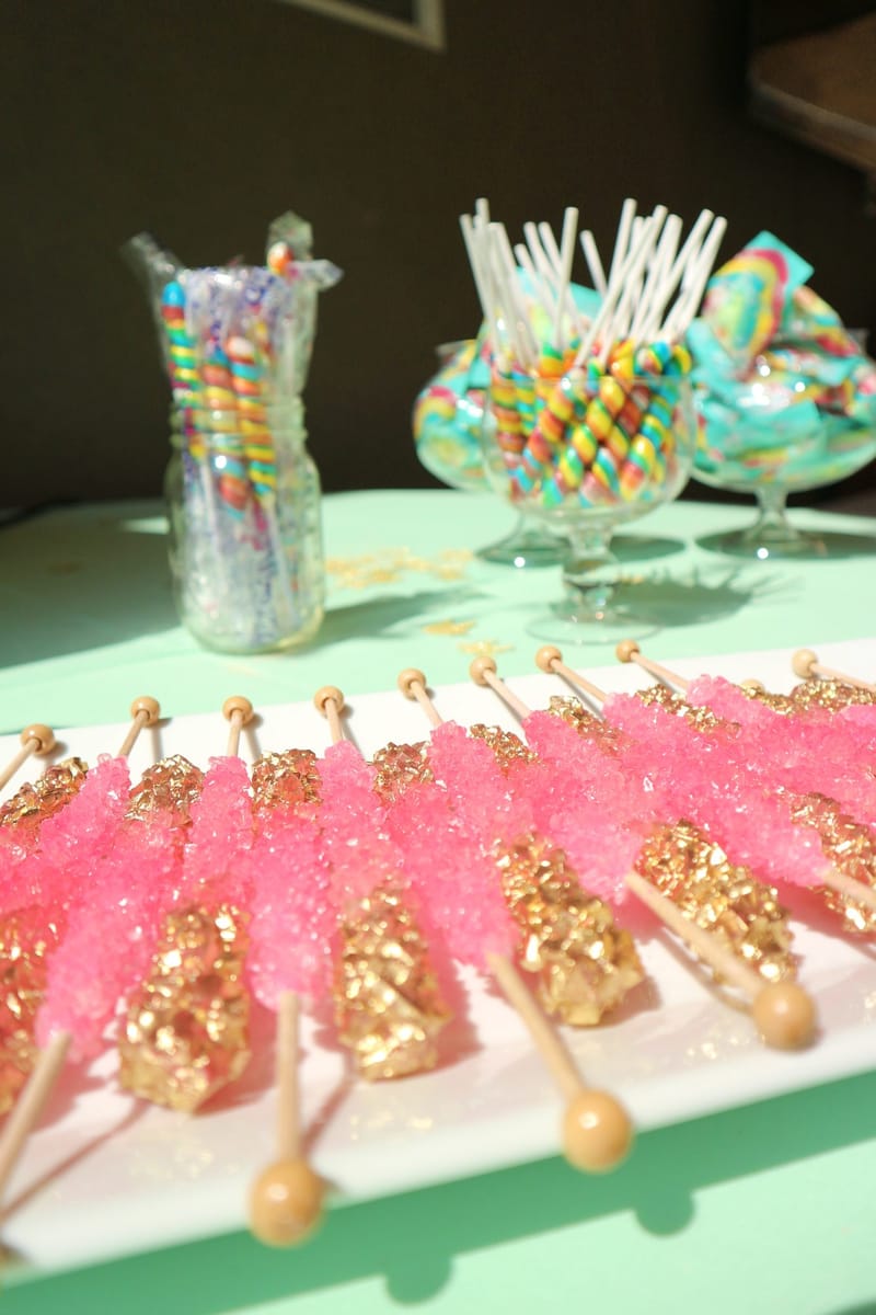 Pink and gold rock candy sticks in a row below jars of rainbow lollipops