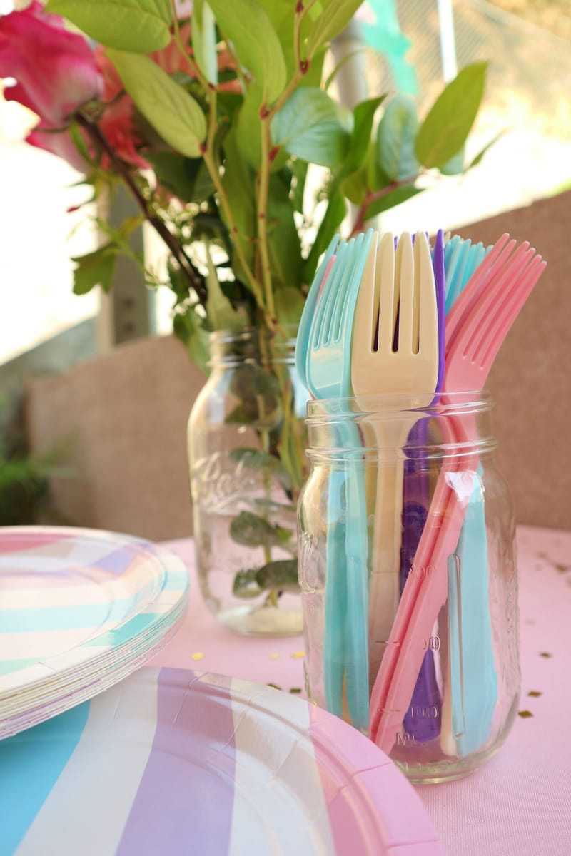 Pastel plastic forks in a glass jar beside a vase of roses and striped paper plates