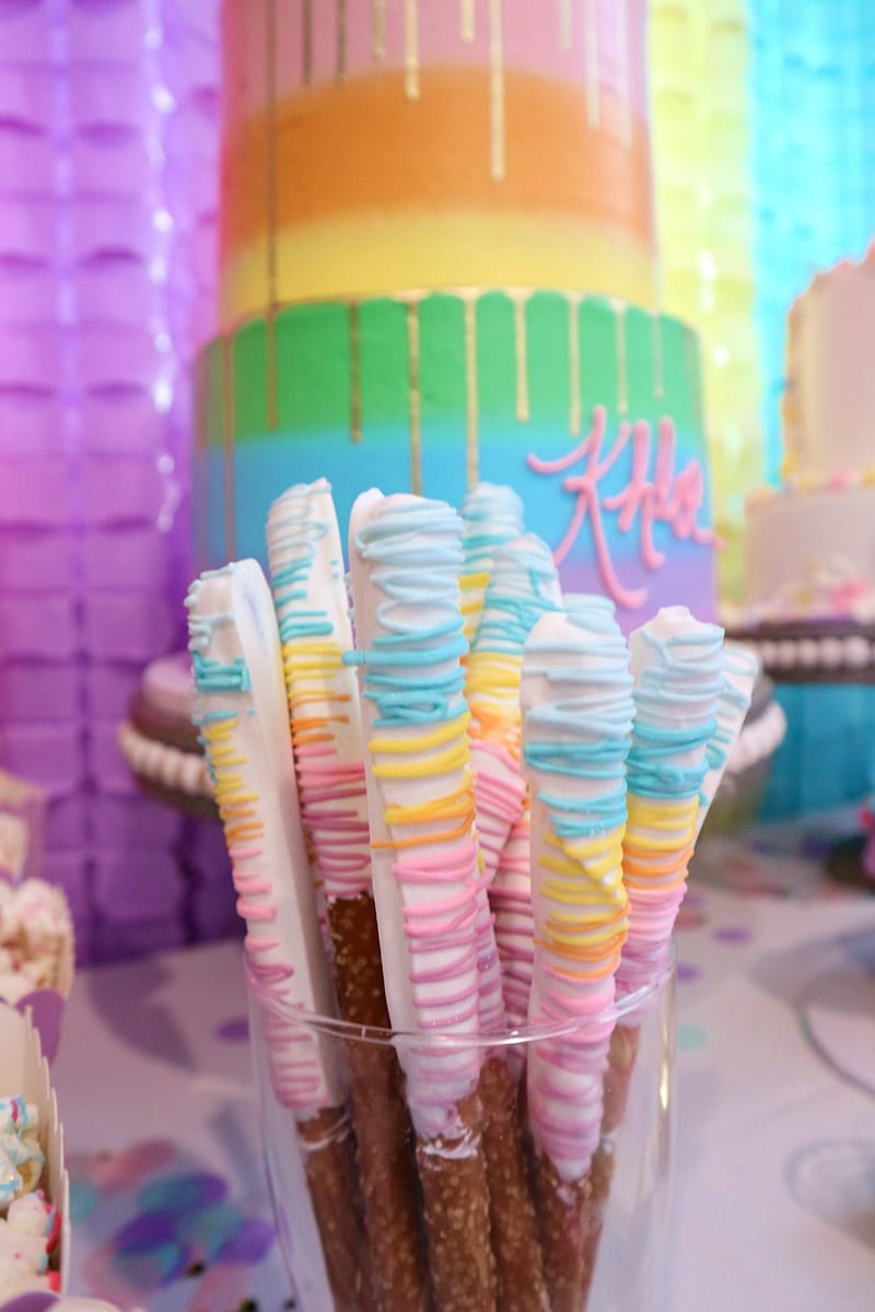 Pretzel rods dipped and drizzled in pastel colors in front of the rainbow Khloe cake