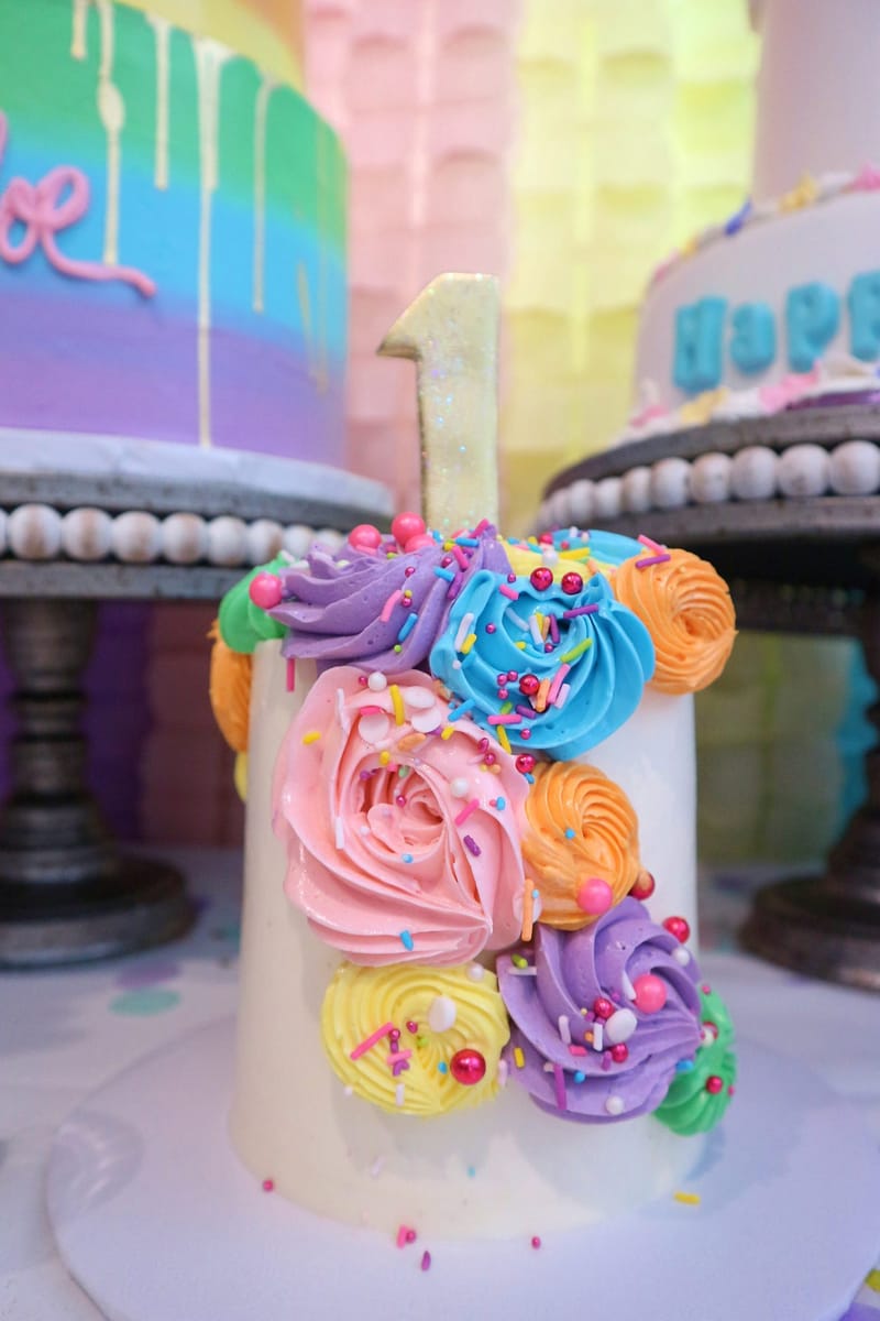 Small white cake with a glitter number 1 topper and swirls of rainbow frosting