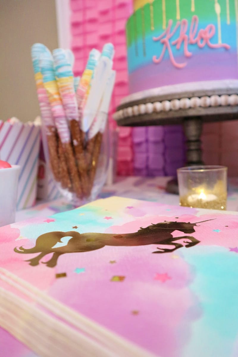 Watercolor napkin with a gold foil unicorn beside dipped pretzel rods, a candle and the rainbow cake