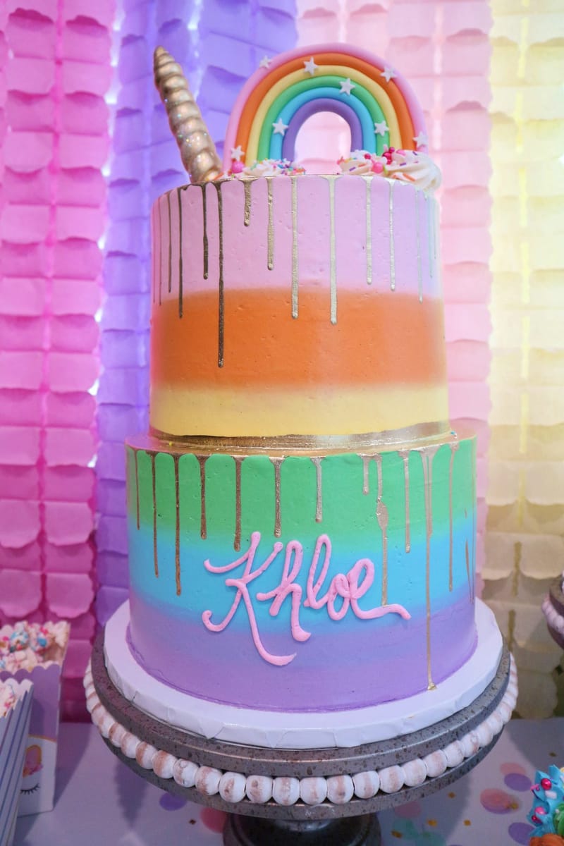 Two-tier rainbow-striped cake with gold drips, Khloe in pink icing, a rainbow topper and a gold horn