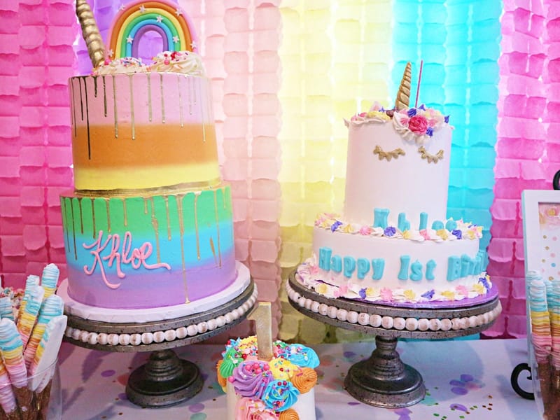 Cake table with the rainbow Khloe cake, the white unicorn cake, a small number 1 cake and dipped pretzels
