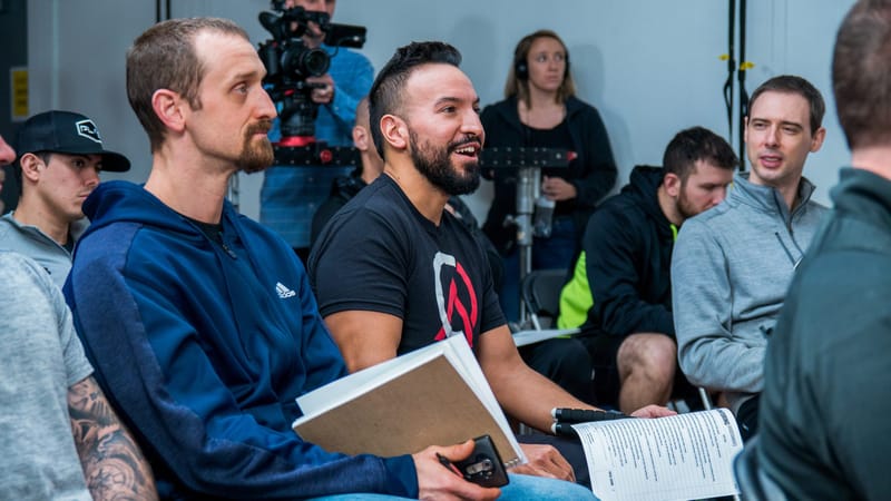 Attendees seated close together with notebooks and handouts, listening, as a video camera records behind them