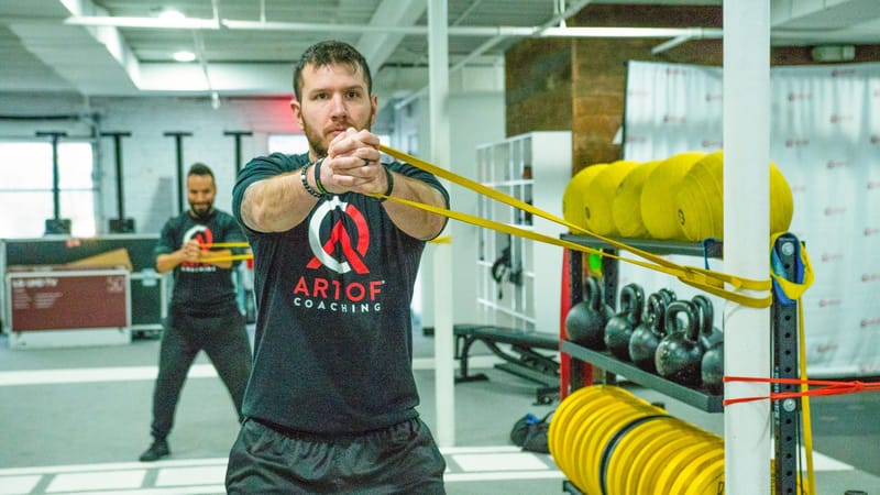Someone stretching a yellow resistance band beside racks of kettlebells and yellow slam balls