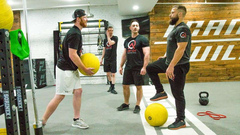 People working through a drill with yellow slam balls on a gym floor before a wood-paneled wall