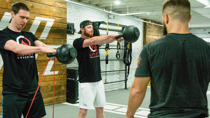 Two people holding kettlebells out at arm's length on a gym floor as another man watches