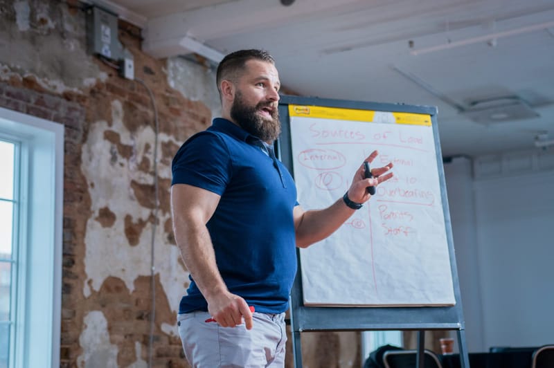The presenter gesturing beside a flip chart of handwritten notes in front of an exposed brick wall