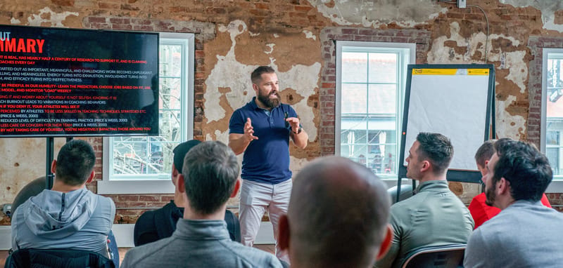 Attendees facing the presenter, a flip chart and a summary slide in a room with brick walls and tall windows