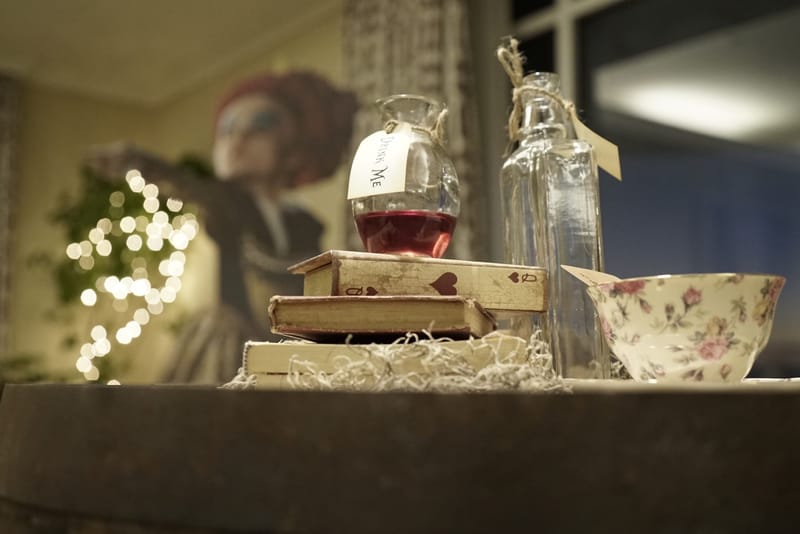 Drink Me bottle of red liquid on stacked books beside a floral bowl, a cutout figure blurred behind