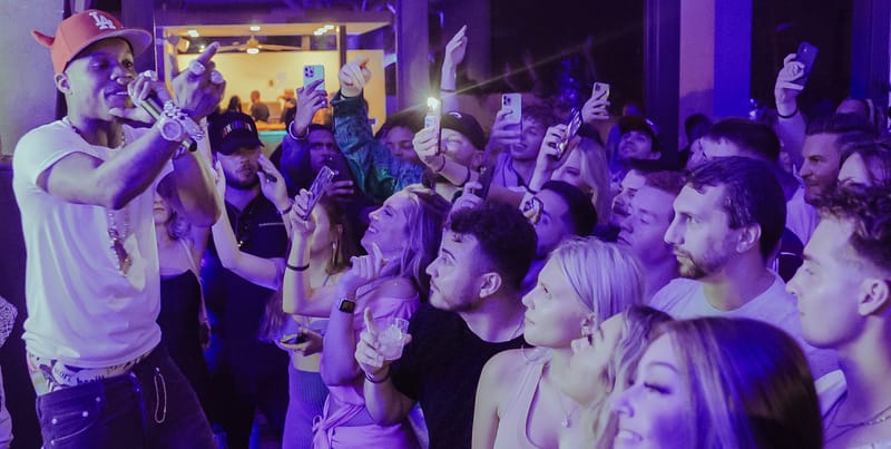 Performer in a white T-shirt with a microphone facing a crowd holding up phones under purple light