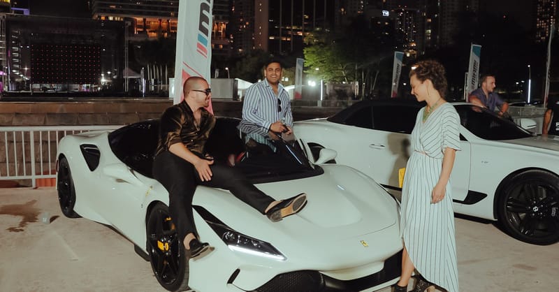 Man sitting on the hood of a white Ferrari at night as two people stand beside it near flags