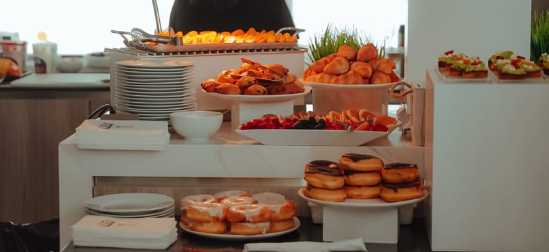 Catering counter with doughnuts, glazed pastries and plates of fruit on white stands, with stacks of plates
