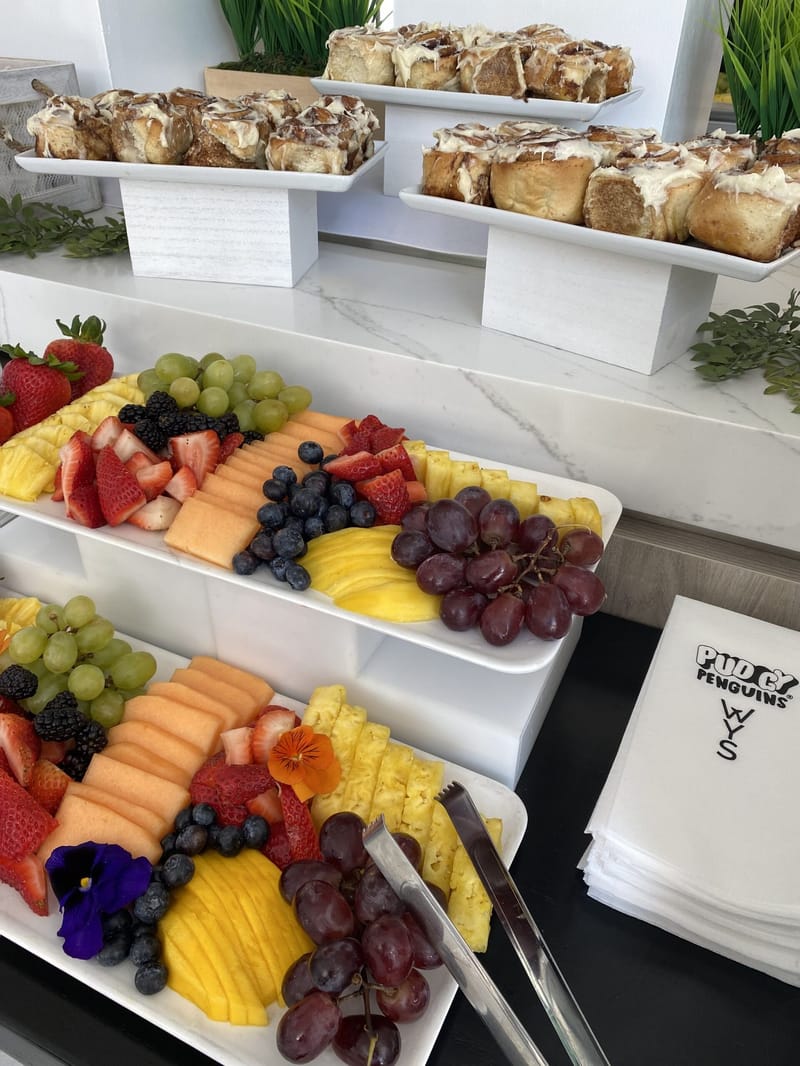 Fruit platters of melon, berries and grapes below iced cinnamon rolls, beside Pudgy Penguins WYS napkins