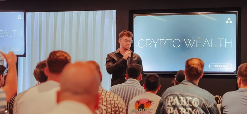 Speaker with a microphone presenting to a seated audience in front of a CRYPTO WEALTH screen