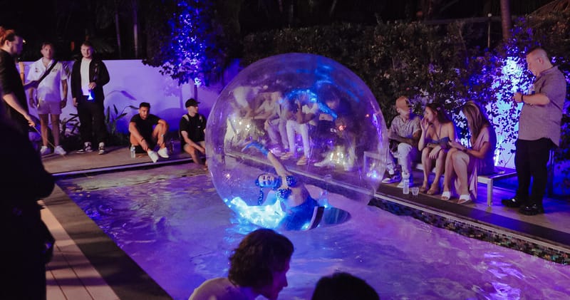 Guest inside a clear inflatable bubble in a pool lit purple at night as people watch from the edge
